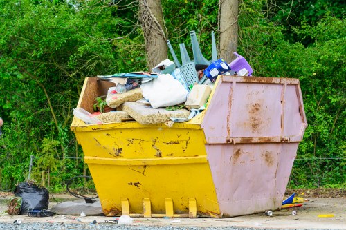 A large open yellow and pink skip positioned on a gravel surface outdoors, filled with various discarded household items including plastic chairs, a white plastic bag, cardboard, and gravel. The skip shows some signs of dirt and rust, with the background featuring green trees and foliage, indicating it is situated in a natural environment near a wooded area. The scene is illuminated by natural daylight, emphasizing the piled rubbish and the texture of the skip's metal surface. This image relates to waste collection and rubbish disposal services that companies like Office Clearance Swiss Cottage may provide in the local London area, such as around Swiss Cottage postcodes.