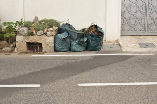 A trio of large dark green refuse sacks filled with rubbish, placed on the edge of a paved street near a white wall with a small garden bed containing green plants and rocks behind them. The refuse sacks have a slightly crumpled texture and are positioned close together, leaning against the wall. To the right, there is a metal drainage grate and a decorative grey metal gate. The street surface is dark asphalt with white painted lines, and the scene appears to be part of a residential or commercial area within or near Swiss Cottage, London, suggesting a location where rubbish removal services by Office Clearance Swiss Cottage may operate to manage waste collection and disposal.
