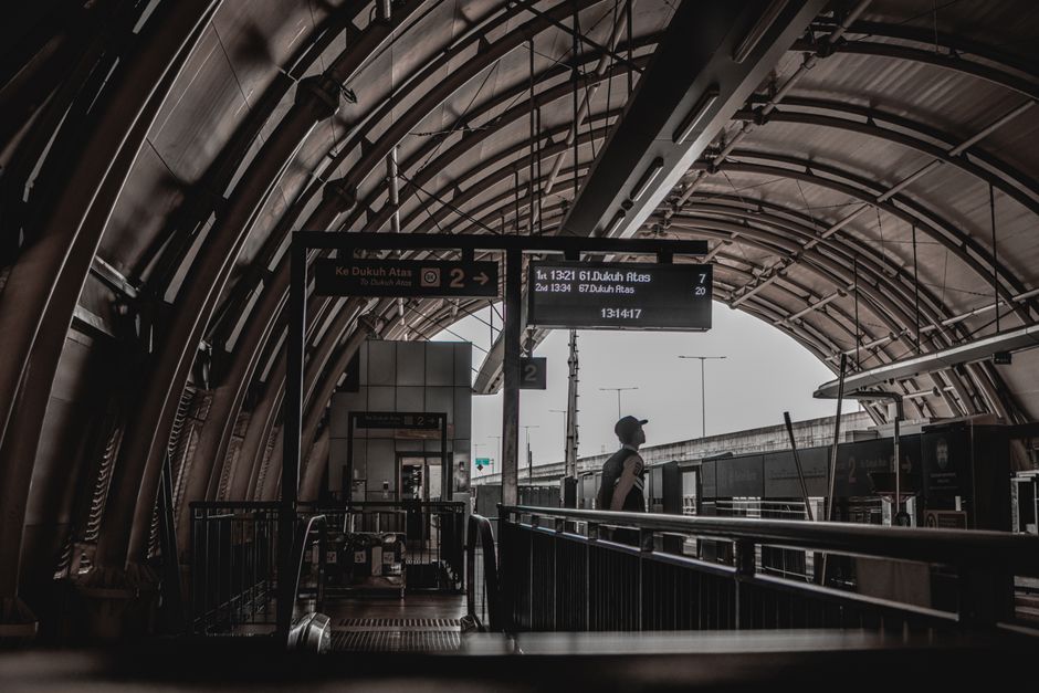 Inside a modern railway station with a curved, metallic arch ceiling supported by visible structural beams. The platform area is illuminated by natural light filtering through large glass panels at th