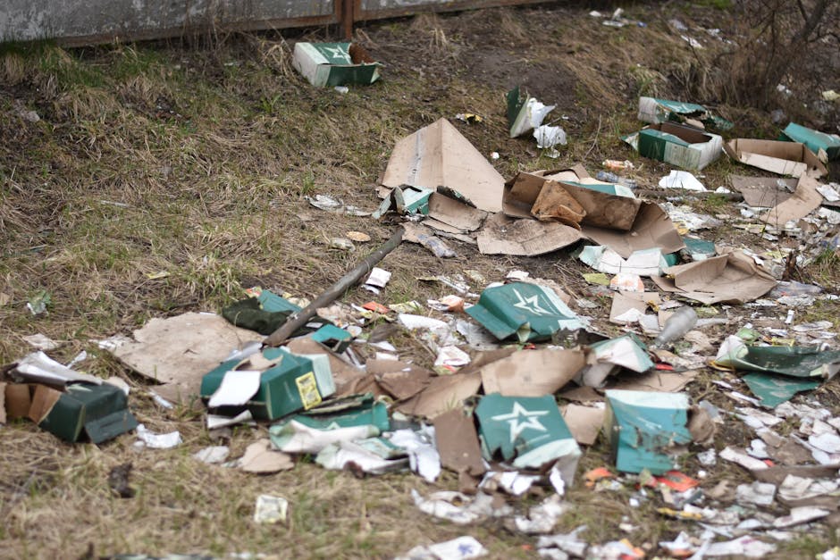 An outdoor scene showing a patch of uneven grassy ground with scattered litter, including flattened and torn cardboard boxes, some green with white star logos, and crumpled pieces of paper and plastic